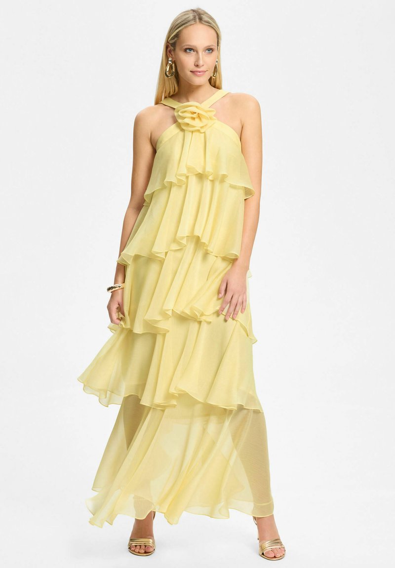 Femme portant une robe dos nu fluide et superposée de couleur jaune pâle avec un détail floral au niveau de l'encolure et des talons hauts dorés à brides.