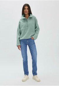 Pull en polaire vert clair avec un demi-zip et un col haut, associé à un jean bleu et des baskets beiges. Coupe décontractée et texture douce.