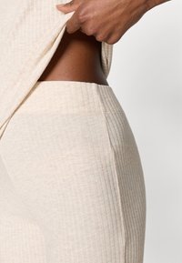 Σετ άνεσης σε μπεζ ριμπ ύφανση που δείχνει την υφή του υφάσματος· το μπλουζάκι σηκώνετα ι αποκαλύπτοντας λεία επιδερμίδα· η ζώνη του παντελονιού είναι ορατή με λεπτομέρειες ελαστικότητας.