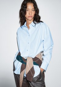 Lichtblauw overhemd met knopen en lange mouwen, gecombineerd met een bruin en blauwgroen trui met ruitpatroon, in de taille geknoopt, gedragen over een grijze broek.