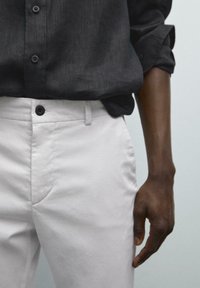 Weiße Baumwollhosen mit glatter Textur, seitlichen Taschen und schwarzem Knopfverschluss, kombiniert mit einem dunkelgrauen Hemd mit Knöpfen.