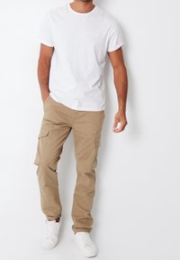 Hvid T-shirt med korte ærmer kombineret med beige cargobukser med sidelommer, båret med hvide sneakers, stående mod en ensfarvet baggrund.