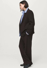 Czarny dopasowany garnitur z usztywnioną marynarką i luźnymi spodniami, nałożony na jasnoniebieską koszulę w paski. Gładka tkanina o wyraźnych liniach.