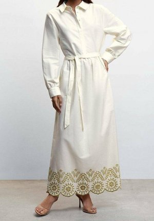 Kobieta ubrana w długą białą sukienkę koszulową z przewiązaniem w talii i misternym kwiatowym haftem na dole, do której dobrała beżowe sandały na wysokim obcasie.