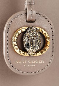 Accessorio in pelle taupe con un lucchetto circolare dorato, un centro in pietra nera testurizzata e il nome del marchio inciso sotto.