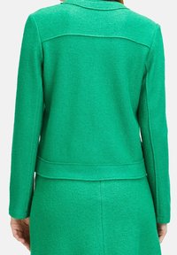 Grön texturerad jacka med långa ärmar, figursydd design och krage. Slät tyg med en subtil lyster, med sömmar längs ryggen.