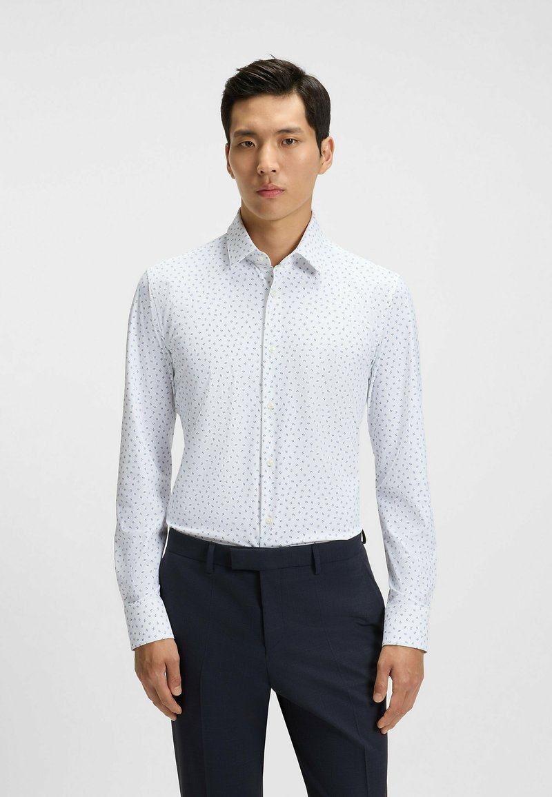 Vit långärmad skjorta med knappstängning och små blåa bladmönster, figursydd passform och en välstruken krage, tillsammans med mörka byxor.