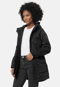 Black parka jacket with a hood, featuring a zip closure, front pockets, and a subtle logo on the sleeve. Worn over a white shirt and dark jeans.