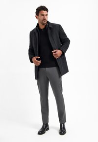 Donkergrijze wolmixjas met een kraag, gedragen over een zwarte polo, gecombineerd met grijze op maat gemaakte broeken en zwarte leren laarzen.