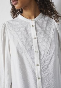 Hvid bluse med høj krave, der har en bølget, broderet bærestykke. Knapperne foran er perlemorsagtige, med lange ærmer.