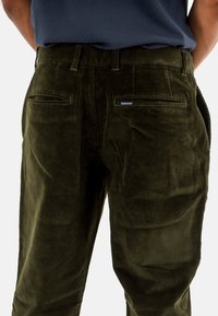 Pantalons en velours côtelé vert olive avec une surface texturée, dotés de deux poches arrière et d'une petite étiquette de marque sur la poche droite.