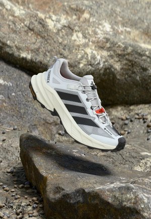 Zapatilla de trail running Adidas Terrex blanca y negra con Gore-Tex, suspendida sobre terreno rocoso.