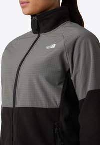 Osoba oblečená v dvoubarevném šedém a černém fleecovém zipovém kabátu s logem The North Face na hrudi, límec zapnutý.