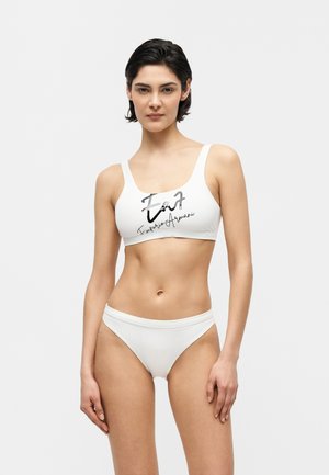 Femeie în picioare purtând costum de baie alb din două piese cu logo-ul „EA7 Emporio Armani” pe partea de sus, pe un fundal alb simplu.