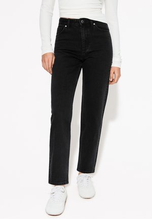 Personne portant un jean noir taille haute à jambes droites, un haut blanc à manches longues et des baskets blanches, debout devant un fond uni.