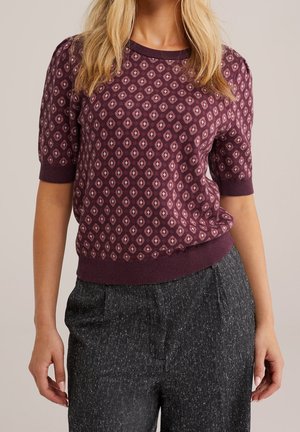 Vrouw draagt een paars kort mouw patroon trui en een hooggetailleerde donkergrijze gespetterde broek, staat met handen ontspannen langs haar lichaam.