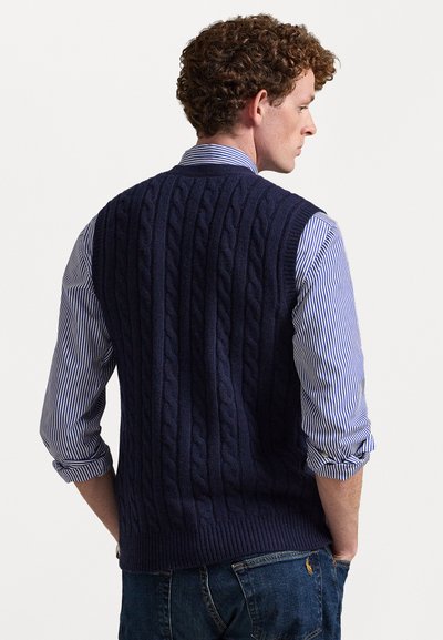 Navy strikket genser vest med kabelmønster, rund hals og ribbestrikket kant. Båret over en blå og hvit stripet skjorte, kombinert med denim jeans.