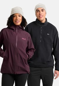 Twee mensen met beige merkbeanies, de een in een kastanjebruine jas met handen in de zakken, de ander in een zwarte fleece pullover.