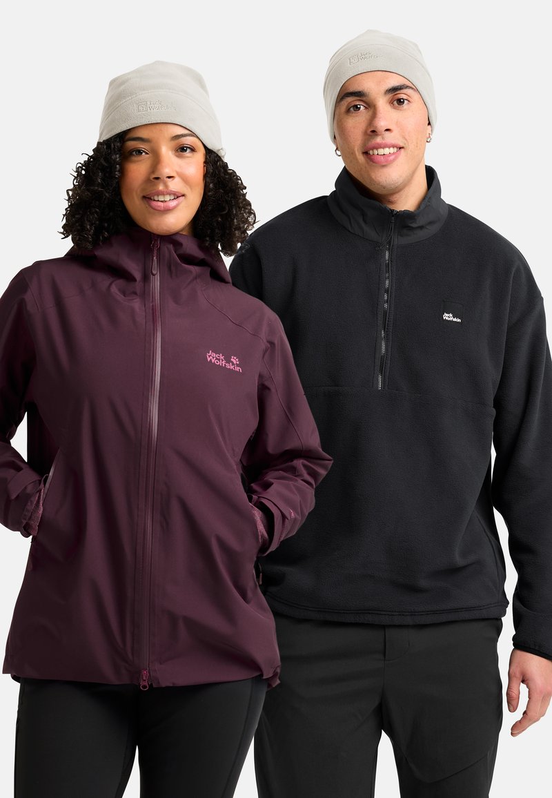 Twee mensen met beige merkbeanies, de een in een kastanjebruine jas met handen in de zakken, de ander in een zwarte fleece pullover.