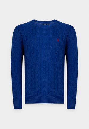 Junger Mann, der einen blauen Zopfpullover über einem karierten Hemd, weite dunkelbraune Hose und weiße Turnschuhe trägt, steht vor weißem Hintergrund.