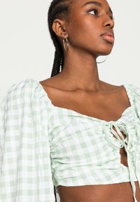 Top cropped em verde e branco xadrez com mangas bufantes, frente com cordão ajustável e tecido texturizado. Brincos de argola prateados visíveis.