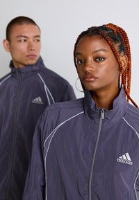 Fiatal nő, aki copfba fonott hajjal és fülbevalókkal áll egy fiatalember előtt; mindketten cipzáros lila Adidas dzsekit viselnek egy sima háttér előtt.
