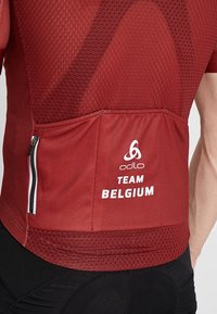 Rød cykeltrøje med mesh struktur, har en baglomme med lynlås og "TEAM BELGIUM" trykt i hvidt. Odlo-logo på den øverste del af ryggen.