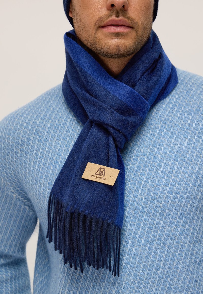 Sciarpa di lana blu navy con bordi frangiati, etichettata "BELLEMERE," indossata sopra un maglione azzurro chiaro con texture.
