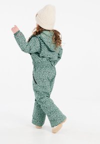 Groene sneeuwoverall met stippen, voorzien van een capuchon, elastische manchetten en ritszakken. Gecombineerd met een beige gebreide muts met textuur en lichtgekleurde laarzen.