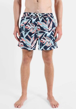 Homme portant un short de bain bleu marine avec un imprimé de feuilles tropicales bleu clair et corail, cordon blanc, debout pieds nus sur un fond uni.