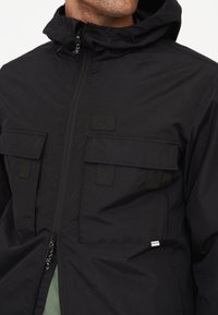 Schwarze Jacke mit einem Reißverschluss vorne, hohem Kragen und zwei Klappentaschen. Hergestellt aus leichtem Material mit einer subtilen Textur und genähten Akzenten.