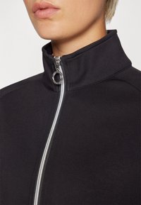 Black zip-up jacket with a high collar, featuring a silver zipper and a circular zipper pull. Smooth fabric with visible stitching details.