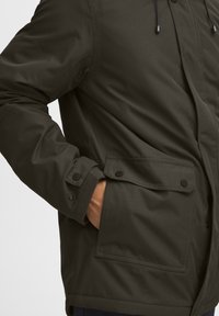 Giacca verde scuro con finitura testurizzata, dotata di zip frontale, tasche con bottone a scatto e cordini regolabili sul cappuccio.