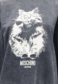 Αφράτη γάτα με γυαλιά και γραβάτα παπιγιόν τυπωμένα σε σκούρο ύφασμα, με το κείμενο «MOSCHINO JEANS» κάτω από την εικόνα της γάτας.