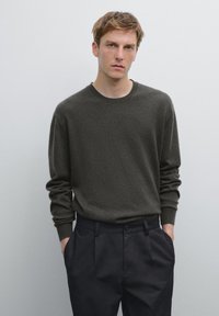 Maglione in lana verde oliva scuro con maniche lunghe e colletto rotondo, abbinato a pantaloni neri con vestibilità morbida e pieghe frontali.