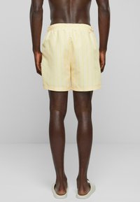 Shorts de bain jaune clair avec des rayures verticales, taille élastique et coupe décontractée, associés à des sandales ouvertes minimalistes.