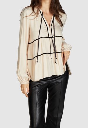 Femme portant un chemisier crème à manches longues avec bordures noires et liens, associé à un pantalon en cuir noir sur un fond uni.