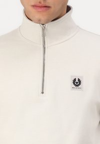 Svetlo béžový pulover na polovičný zips vyrobený z mäkkej textílie. Obsahuje golier, strieborný zips a štvorcový logo nášivku na ľavej hrudi.