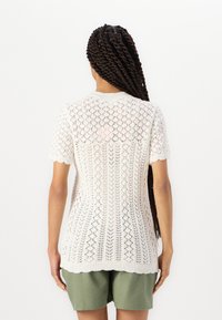 Maglione leggero bianco in maglia con maniche corte, caratterizzato da un motivo in lavorazione a rete e orlo a coste. Il modello indossa pantaloni corti verde oliva.