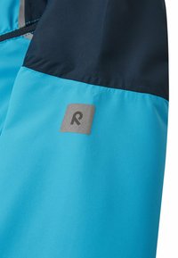 Giacca da esterno blu con una sezione superiore blu navy, texture liscia e logo argento sulla manica. Dettagli delle cuciture a contrasto visibili.