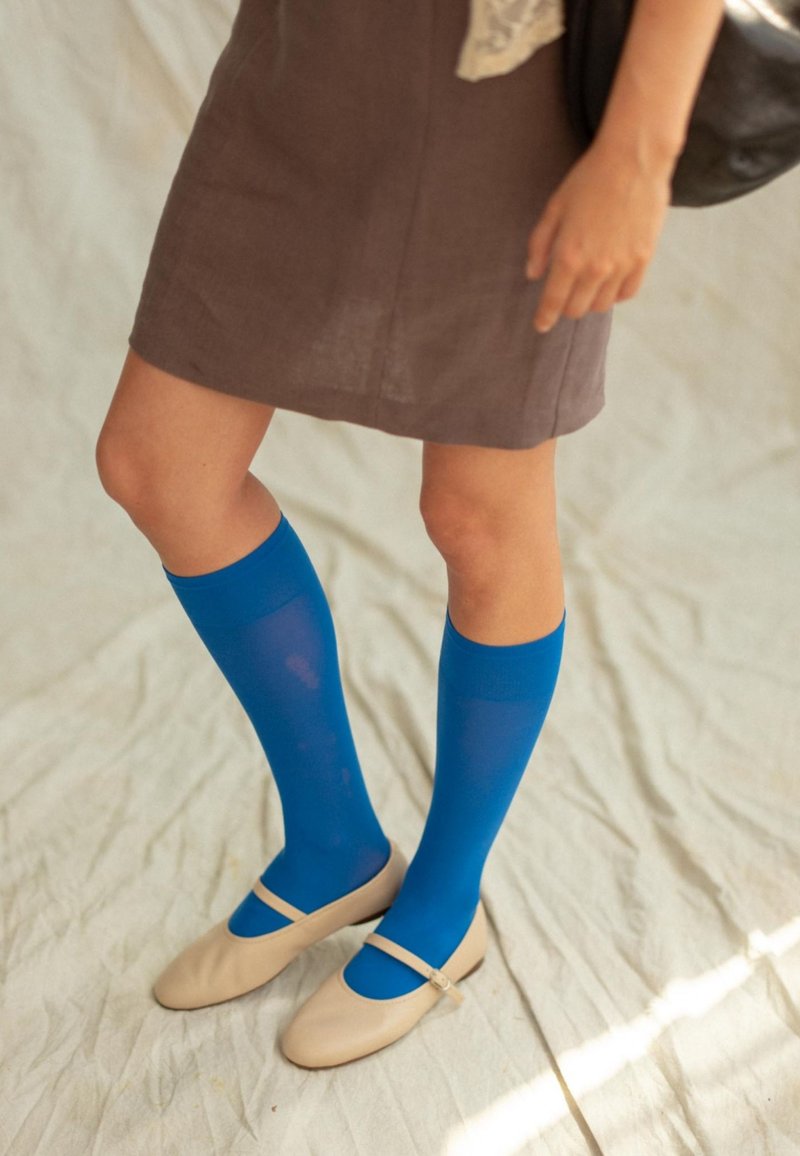 Calcetines azules hasta la rodilla de tela suave combinados con unos zapatos planos beige con tira. El conjunto se complementa con un vestido marrón.