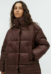 Giacca in stile puffer marrone con colletto alto, silhouette squadrata, sezioni trapuntate e tasche laterali. Texture liscia e lucida.
