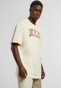 Cream-colored cotton T-shirt featuring bold brown lettering reading "KANYE" and an accent logo, with a relaxed fit and short sleeves.