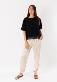 Top corto nero a maniche corte con orlo trasparente, abbinato a pantaloni beige a sigaretta e sandali marroni. L'outfit presenta una vestibilità comoda e tessuti morbidi.