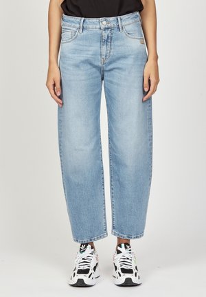 Persoon die lichtblauwe jeans met rechte pijpen en zwart-witte Adidas sneakers draagt, staand tegen een effen achtergrond.