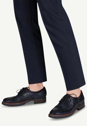 Chaussures en cuir bleu marine de style brogue avec lacets, portées avec un pantalon sombre sur mesure, longueur cheville, sur fond clair.