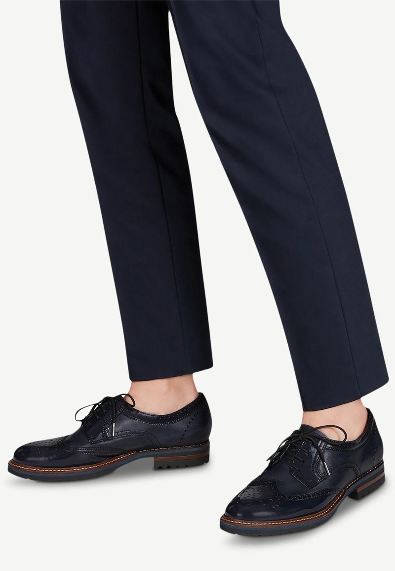 Chaussures en cuir bleu marine de style brogue avec lacets, portées avec un pantalon sombre sur mesure, longueur cheville, sur fond clair.