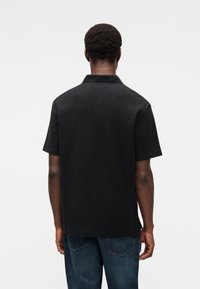 Homme aux cheveux courts et bouclés, portant un polo noir uni à manches courtes et un jean bleu, vu de dos sur un fond blanc.
