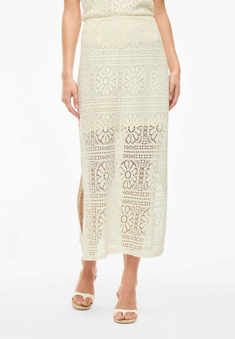 Jupe midi en dentelle beige clair avec des motifs floraux ajourés, dotée d'une fente latérale et d'une texture lisse. Associée à des sandales beiges.