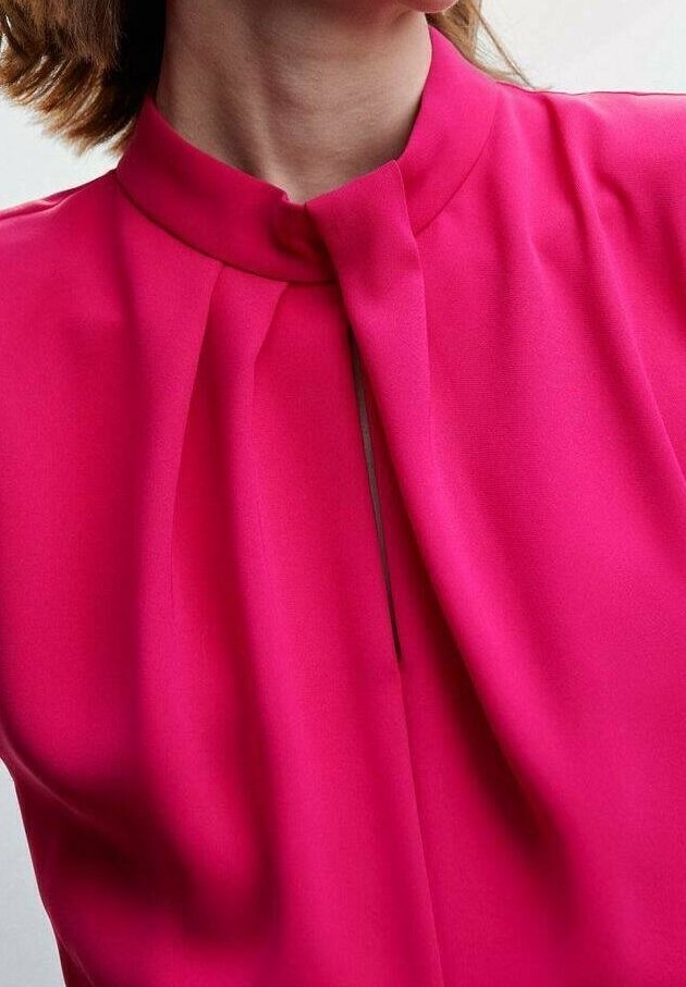 Femme portant un chemisier rose vif avec un col haut et des plis près de l'encolure, montrant une partie de son cou et de ses cheveux.